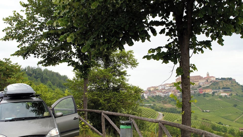 A car parked by a wooden fence overlooking a vineyard and a historic hilltop town.