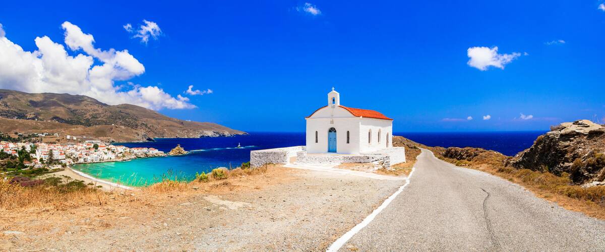 A white church with a red roof on a coastal road, overlooking a turquoise sea and a village.