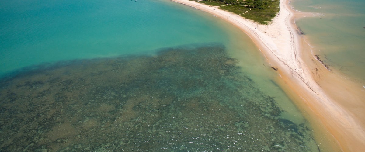 A coastal landscape with a sandy beach, clear turquoise waters, and a small settlement with buildings and greenery.