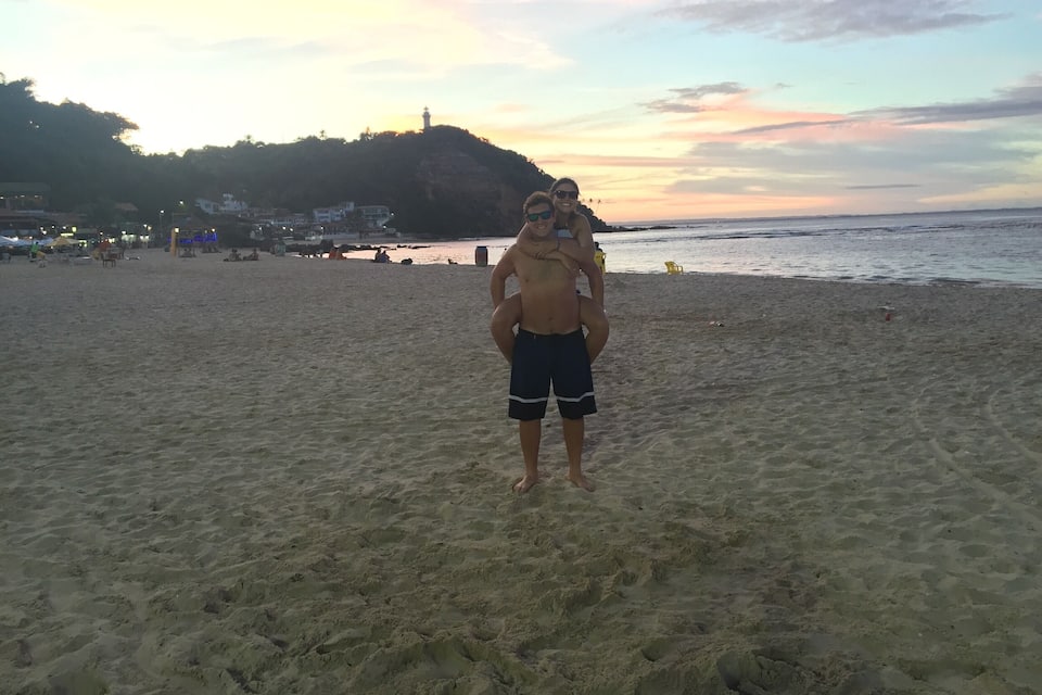 A beach with a person carrying another person on their back, a lighthouse in the background, and a sunset sky.