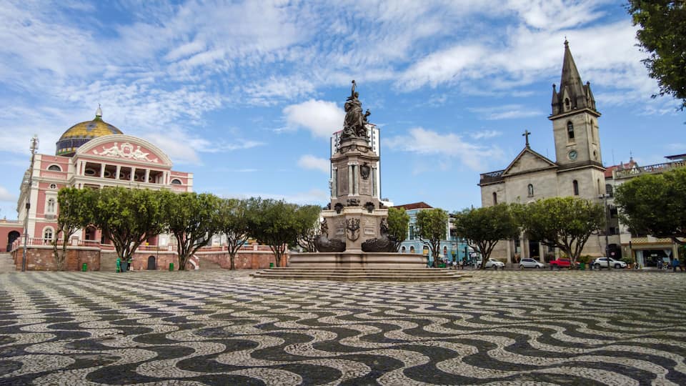 A plaza with patterned cobblestone flooring, a central monument, a church with a tall steeple, and a building with a dome.