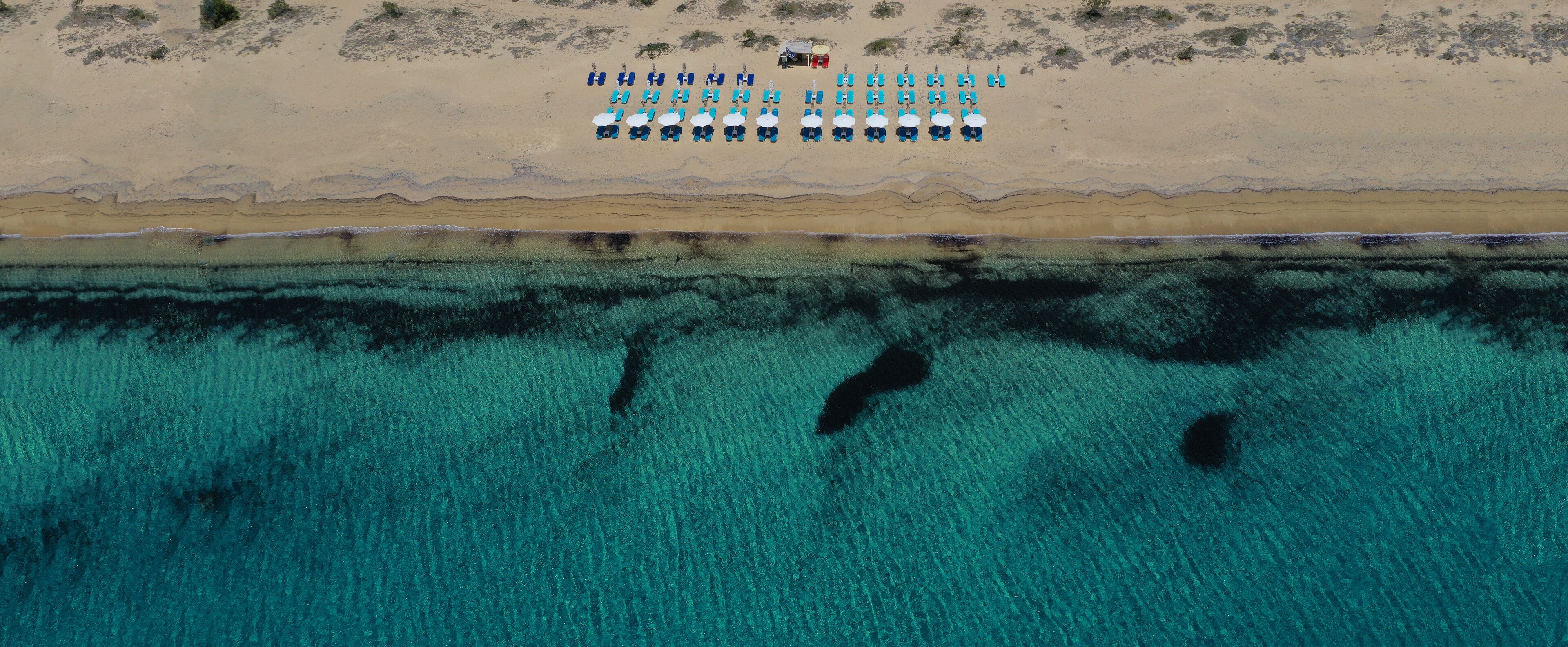 An aerial view of a beach with rows of blue chairs and umbrellas, a small structure on the sand, and clear turquoise water with visible underwater features.