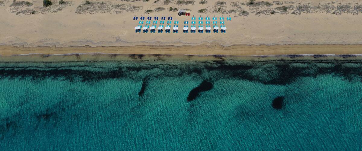 An aerial view of a beach with rows of blue chairs and umbrellas, a small structure on the sand, and clear turquoise water with visible underwater features.