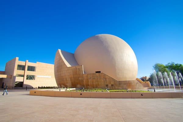 Une structure architecturale moderne dotée d’un grand dôme sphérique, entourée d’une place agrémentée de fontaines et de verdure.