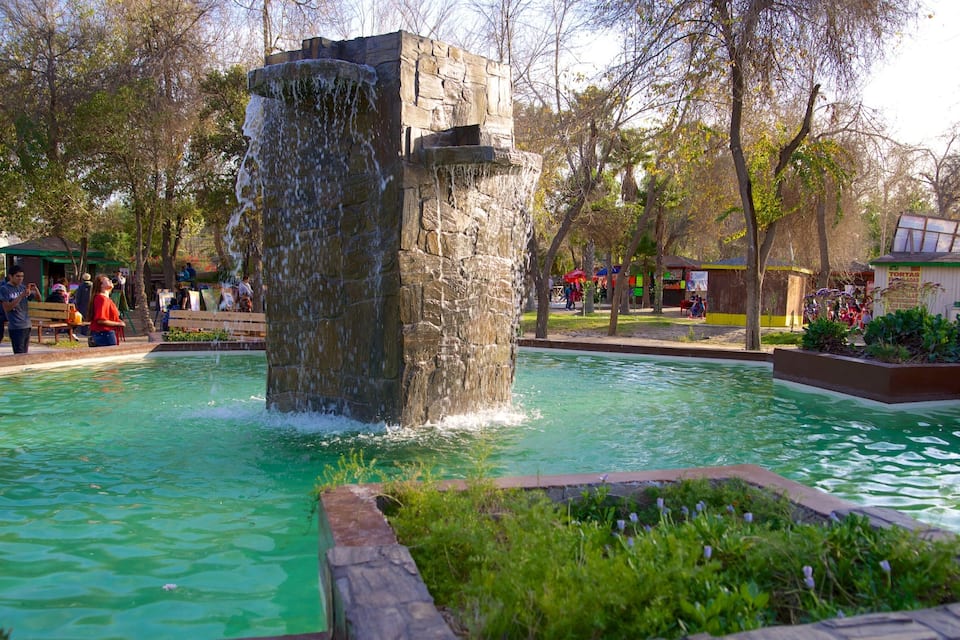 Una fuente de piedra con agua en cascada, rodeada por una alberca y follaje.