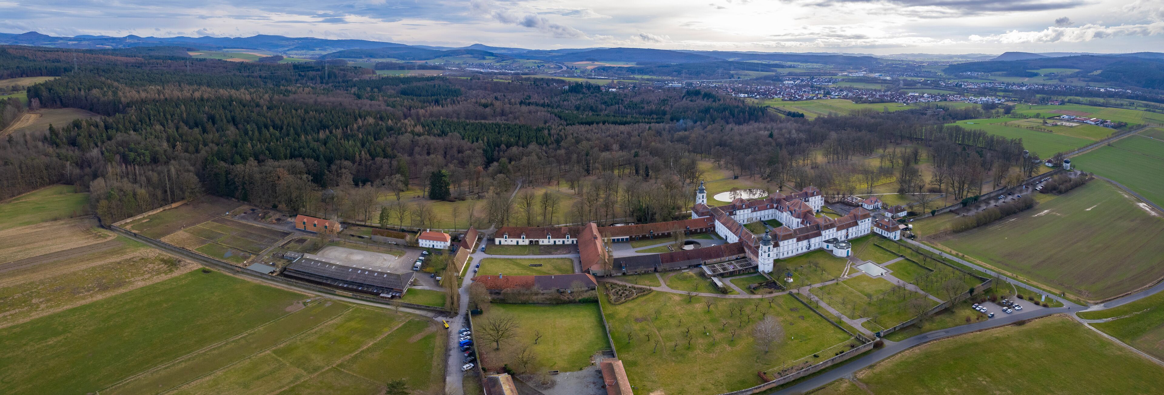 An aerial view of a large estate with expansive green fields, a central building complex, and surrounding forested areas.