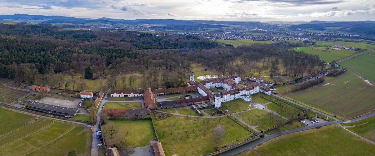An aerial view of a large estate with expansive green fields, a central building complex, and surrounding forested areas.