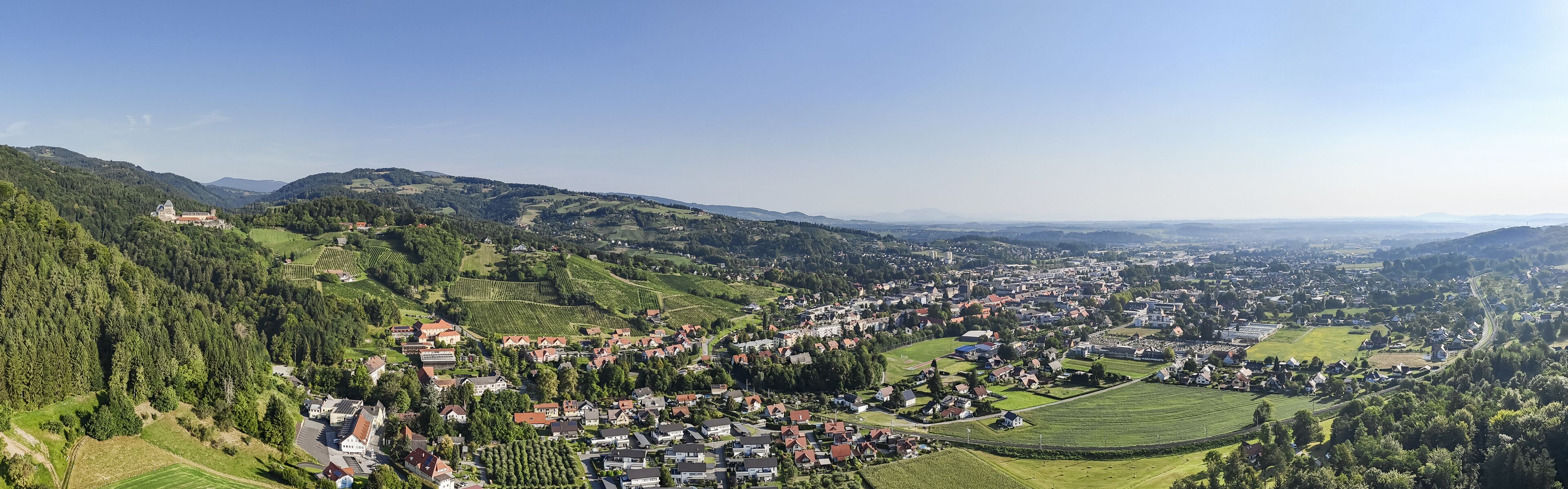 Aerial view of the verdant landscape with the scattered red-roofed buildings and the distant silhouette of a monastery on the hill, Deutschlandsberg, Styria, Austria.