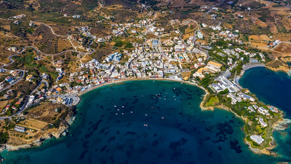 An aerial view of a coastal town with a dense cluster of buildings, a winding road, and a large body of water with boats.