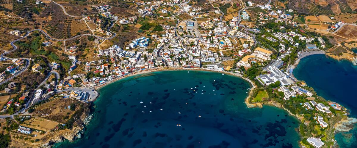 An aerial view of a coastal town with a dense cluster of buildings, a winding road, and a large body of water with boats.