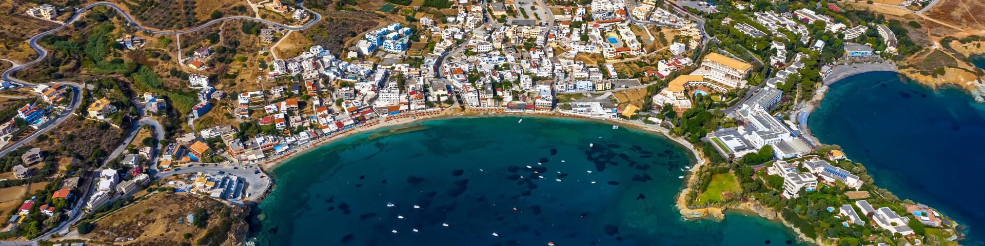 An aerial view of a coastal town with a dense cluster of buildings, a winding road, and a large body of water with boats.