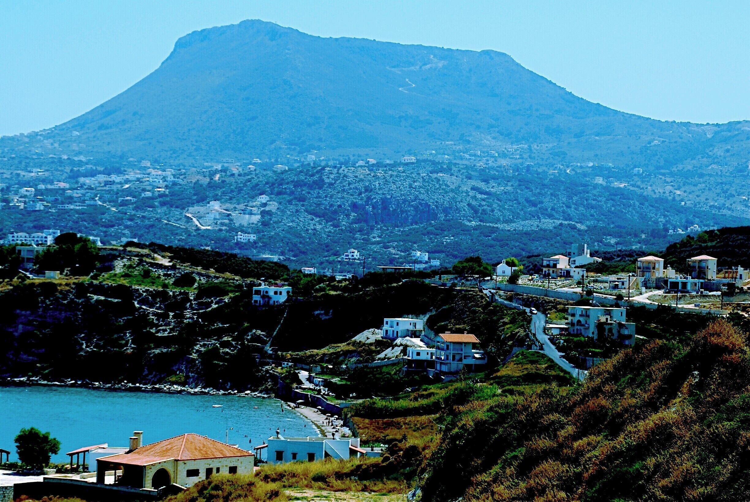 A coastal town with buildings on a hillside, a mountain in the background, and a body of water in the foreground.