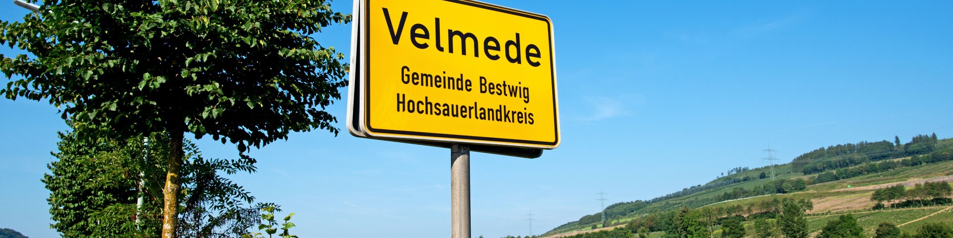 Una señal amarilla con el texto 'Velmede Gemeinde Bestwig Hochsauerlandkreis' sobre un fondo de vía de tren, vegetación y un cielo azul despejado.