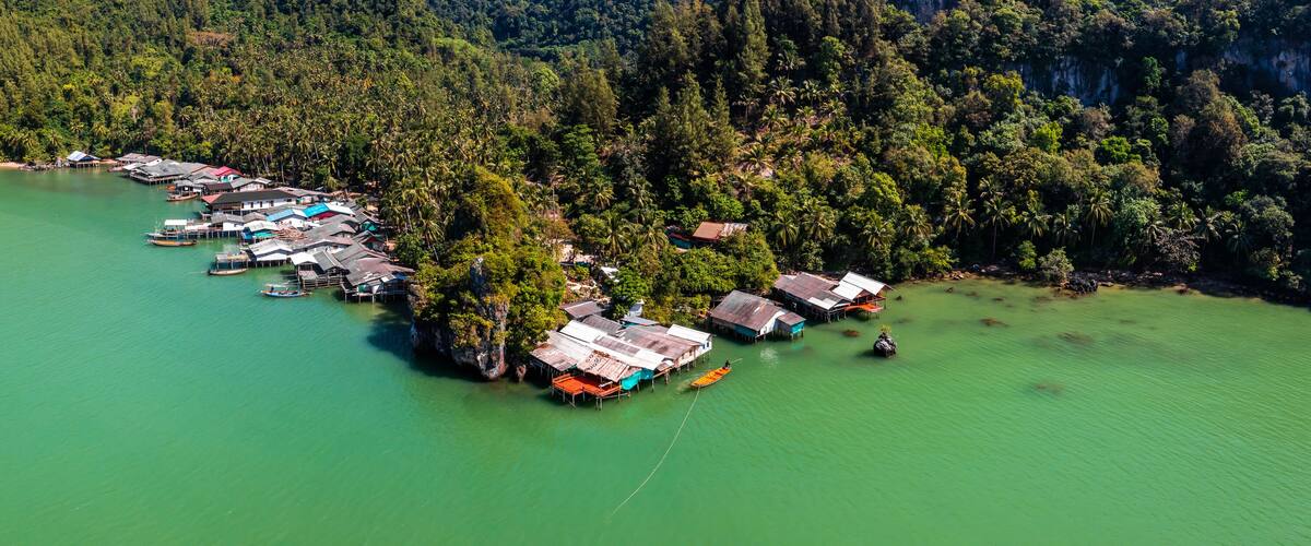 An aerial view of a coastal village with floating houses connected by walkways over calm green waters, surrounded by dense forest and hills.