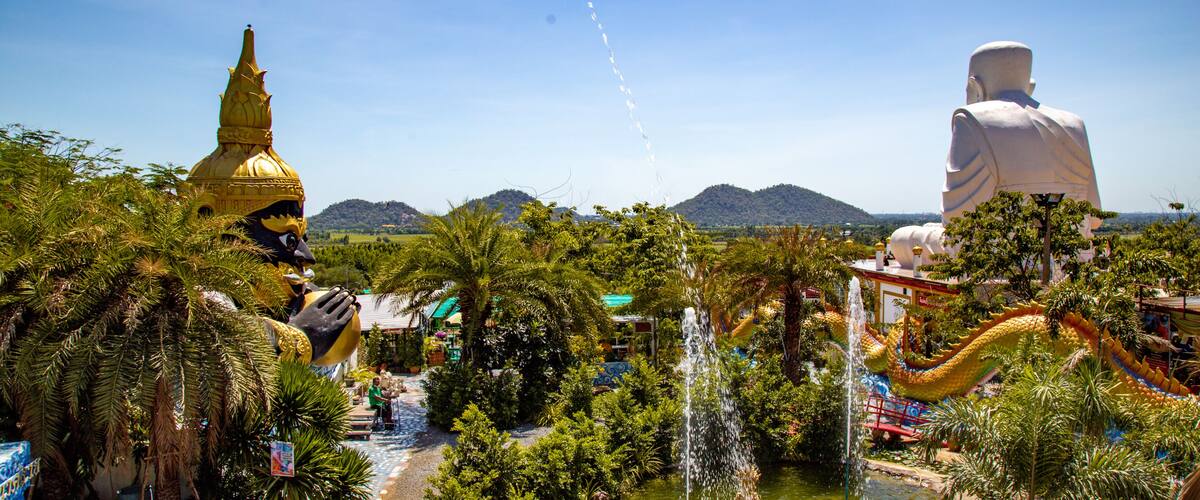 A large golden stupa, a white statue, and a dragon sculpture in a lush garden with mountains in the background.
