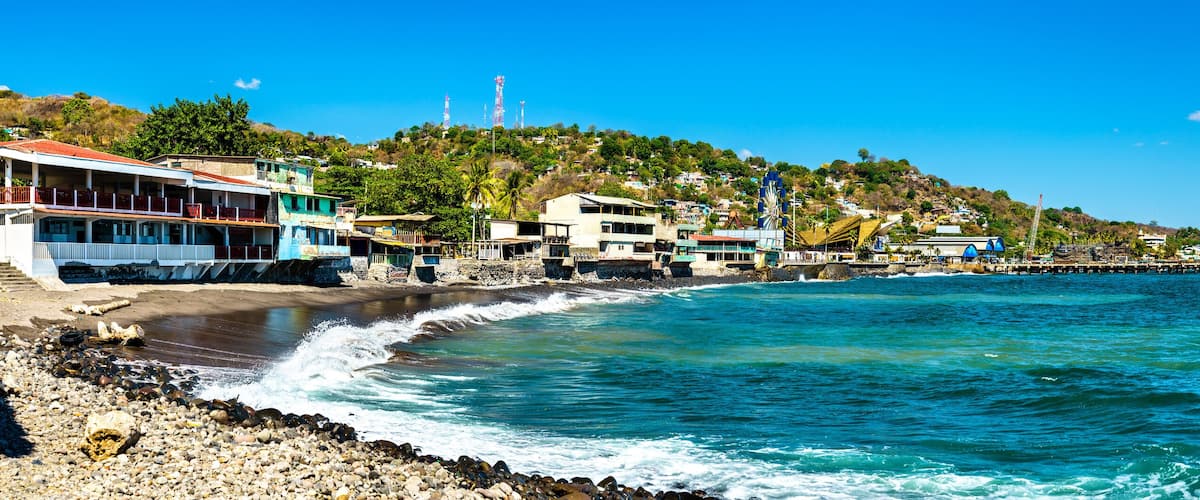 A coastal town with buildings along the shore, a pier extending into the sea, and hills in the background.