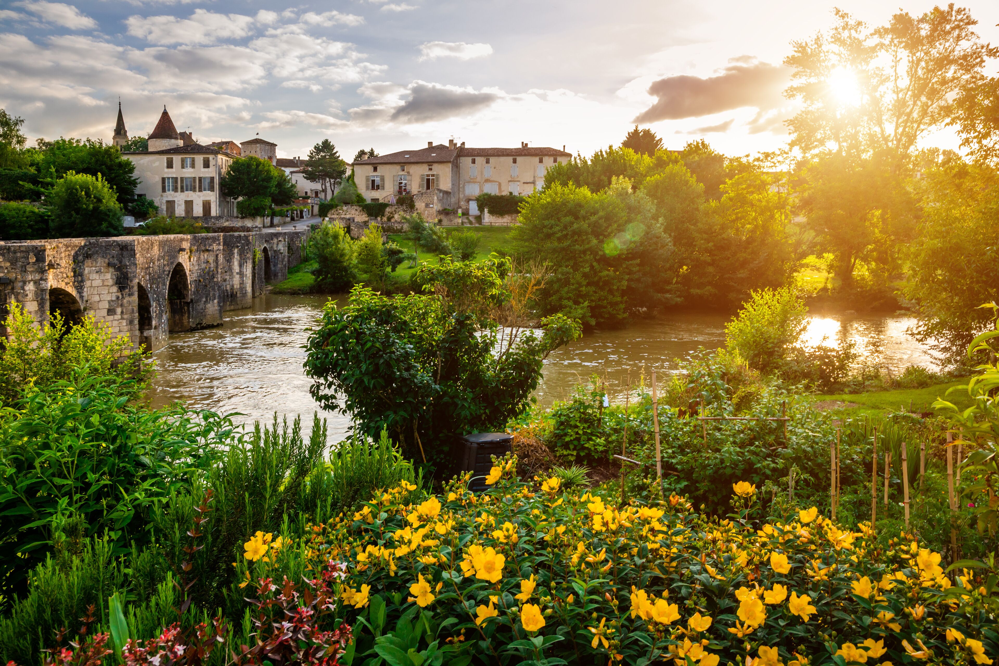 A stone bridge over a river, surrounded by lush greenery and yellow flowers. In the background, there are historic buildings with a church steeple.