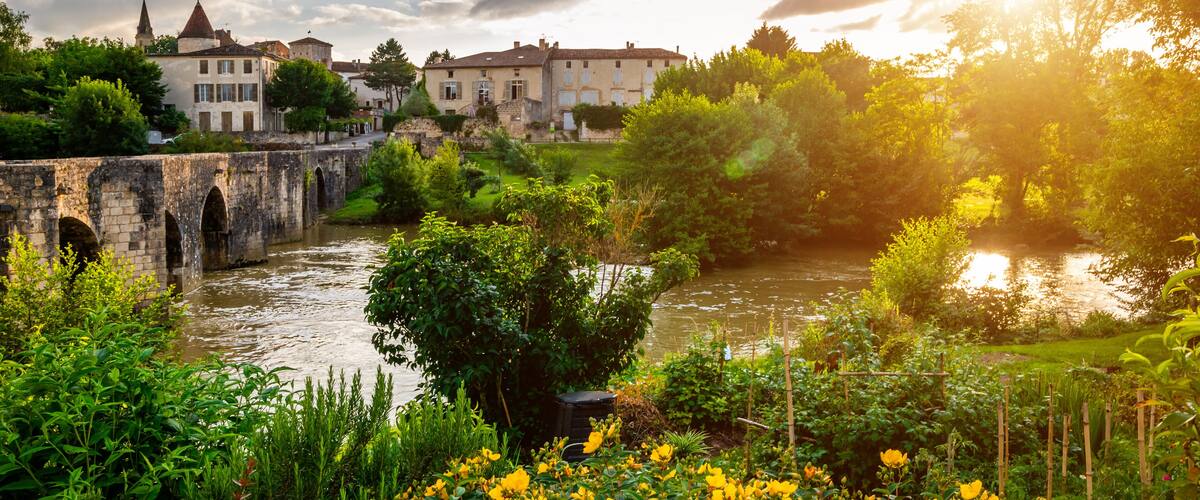 A stone bridge over a river, surrounded by lush greenery and yellow flowers. In the background, there are historic buildings with a church steeple.