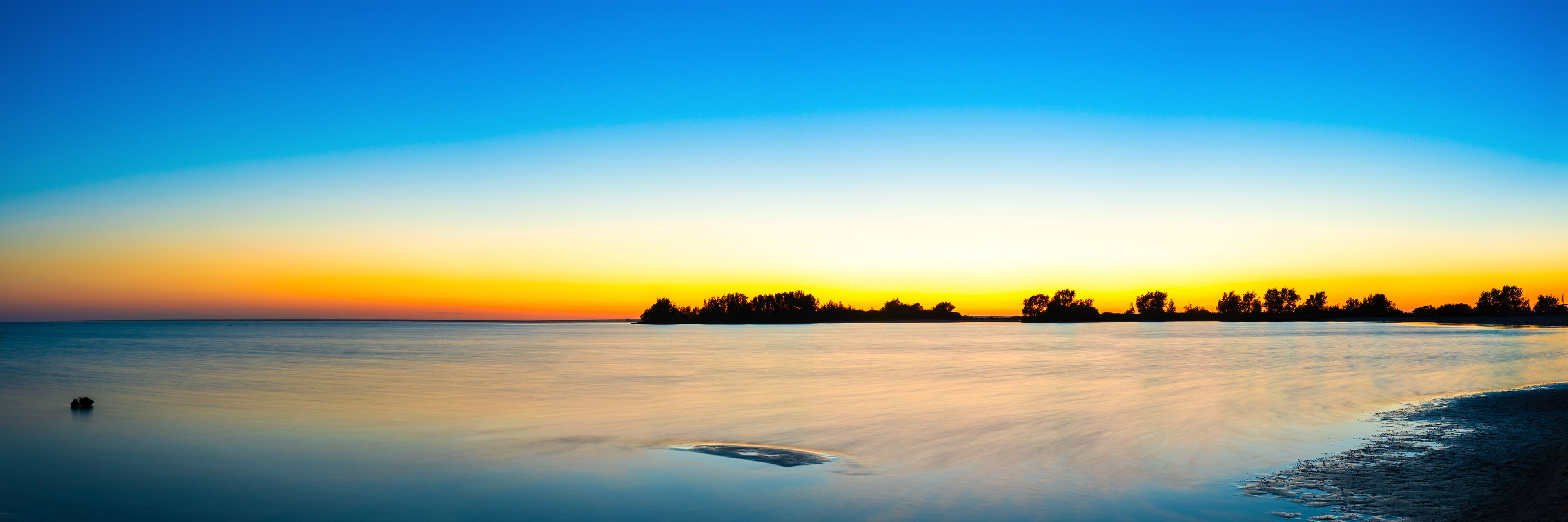 A calm lake at sunset with a silhouette of trees on the horizon and a small boat in the water.