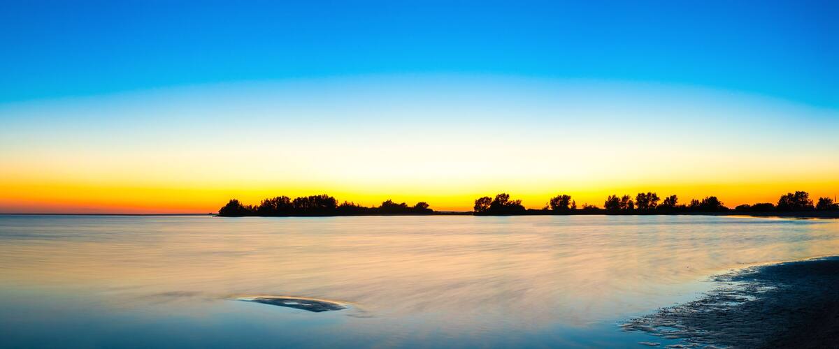 A calm lake at sunset with a silhouette of trees on the horizon and a small boat in the water.