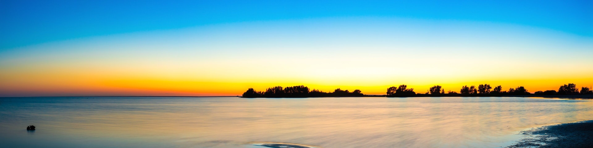 A calm lake at sunset with a silhouette of trees on the horizon and a small boat in the water.