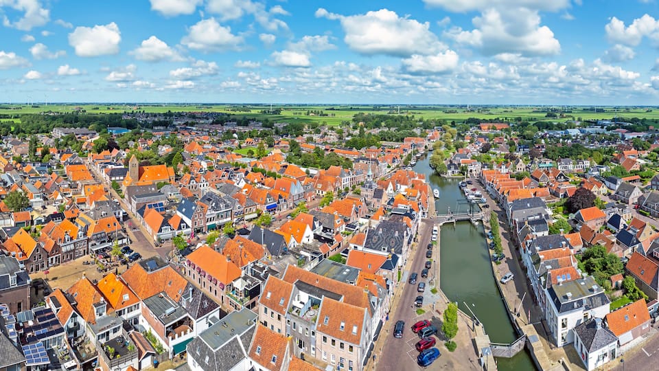 A town with red-roofed buildings, a river, and wind turbines in the distance.