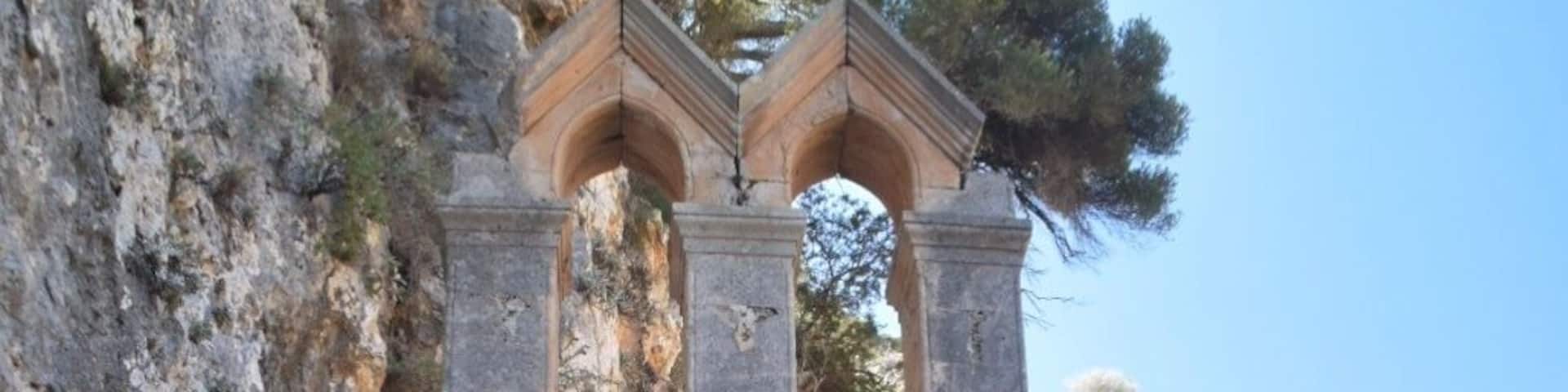 An ancient stone structure with a central doorway and two arches on top, surrounded by rocky terrain and sparse vegetation.