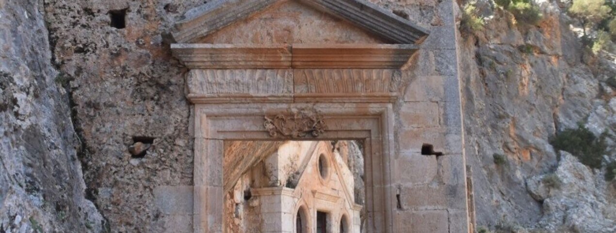 Una antigua estructura de piedra con un acceso central y dos arcos en la parte superior, rodeada de terreno rocoso y vegetación escasa.