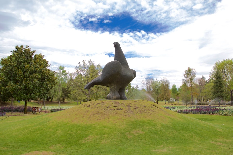 Uma escultura de um animal grande numa colina gramada, com árvores e flores ao fundo.
