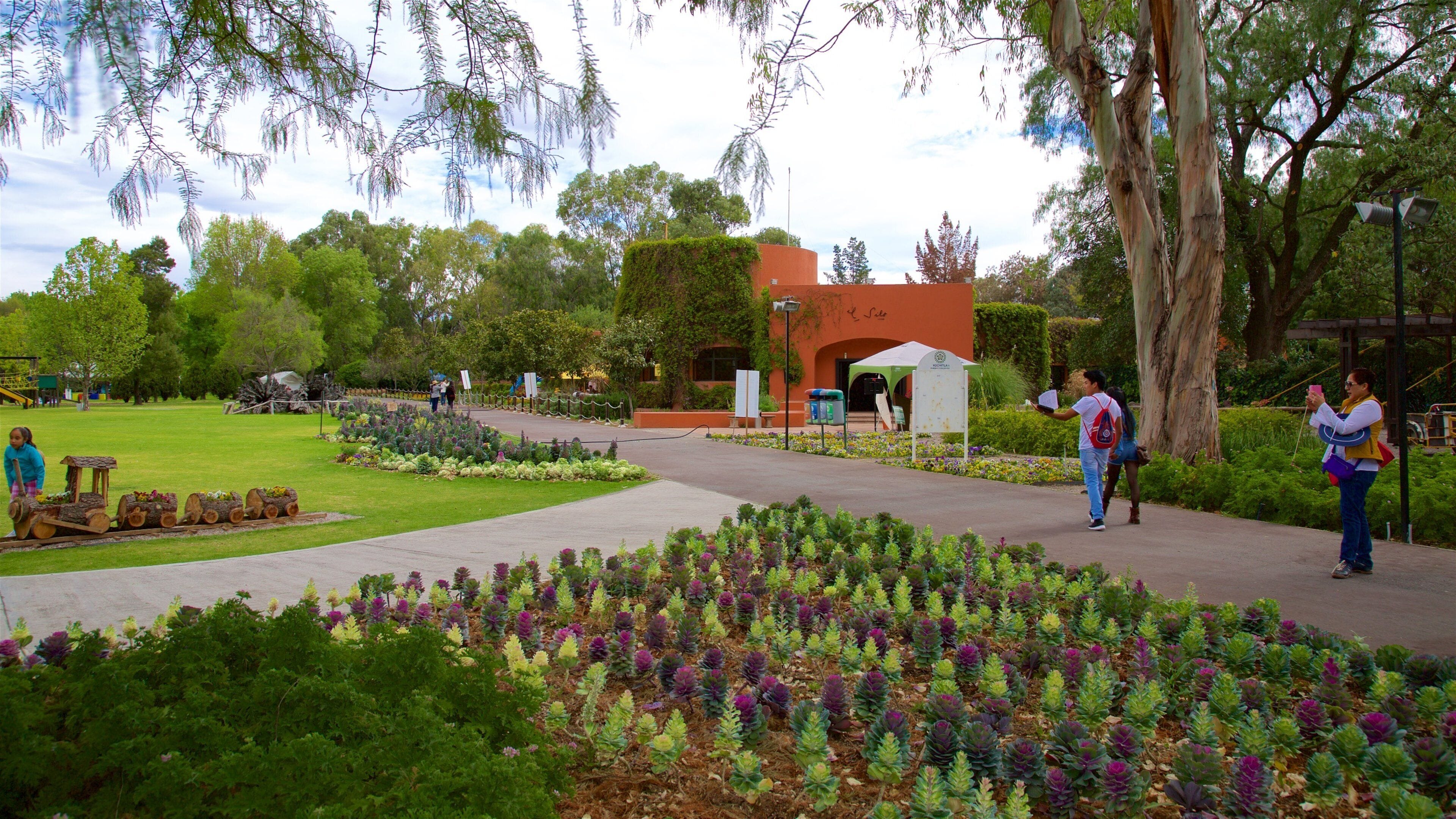 A park with a train track, colorful flower beds, and a building with an archway.