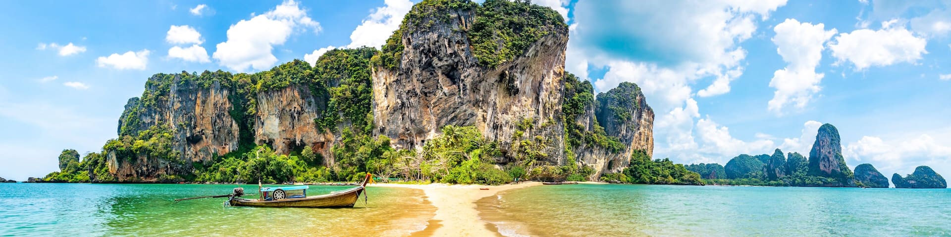 A beach with clear turquoise water, a traditional long-tail boat, and limestone cliffs.