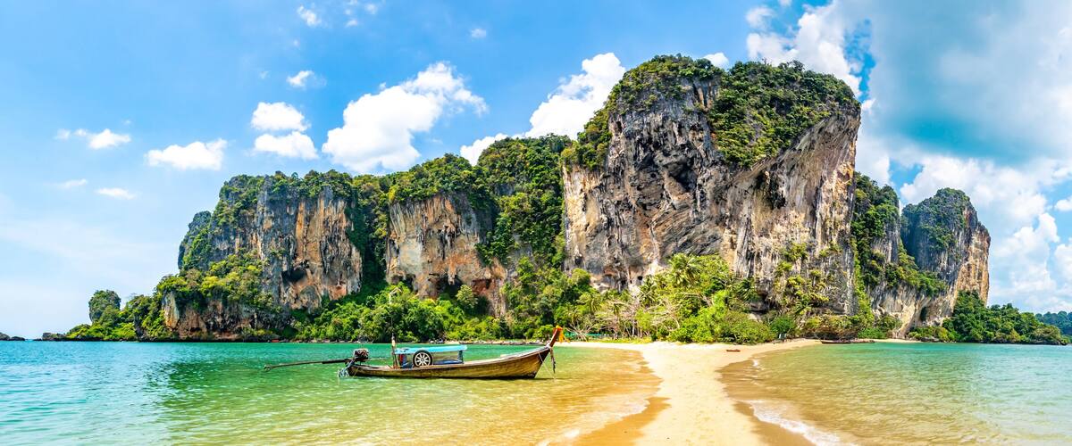 A beach with clear turquoise water, a traditional long-tail boat, and limestone cliffs.