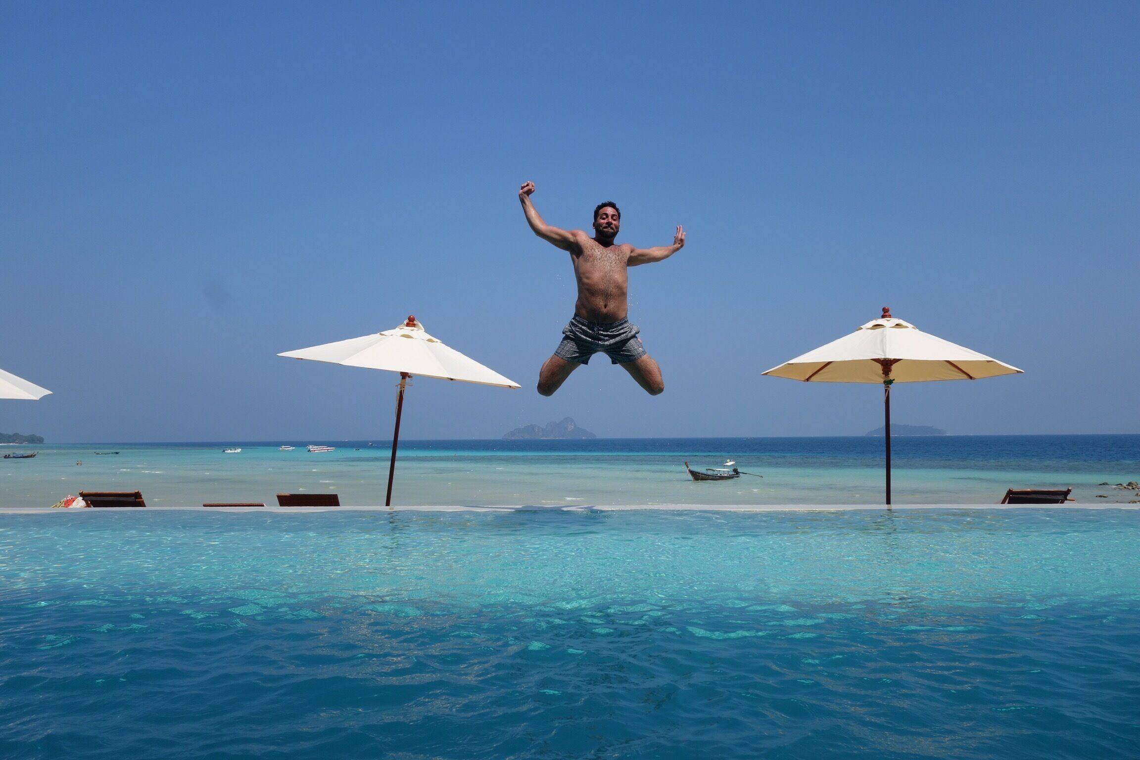 A person jumping into a pool with umbrellas and a clear blue sky.