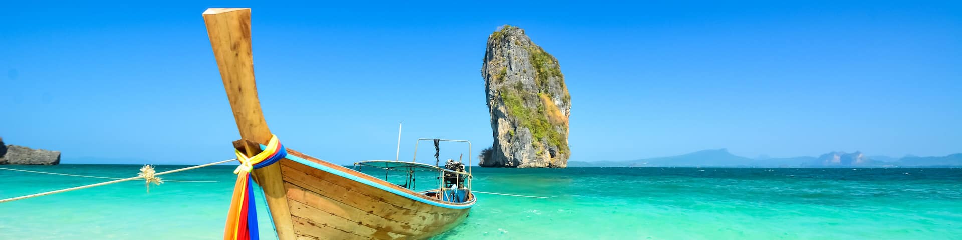 A wooden boat tied to a dock with clear turquoise water and a large rock formation in the background.