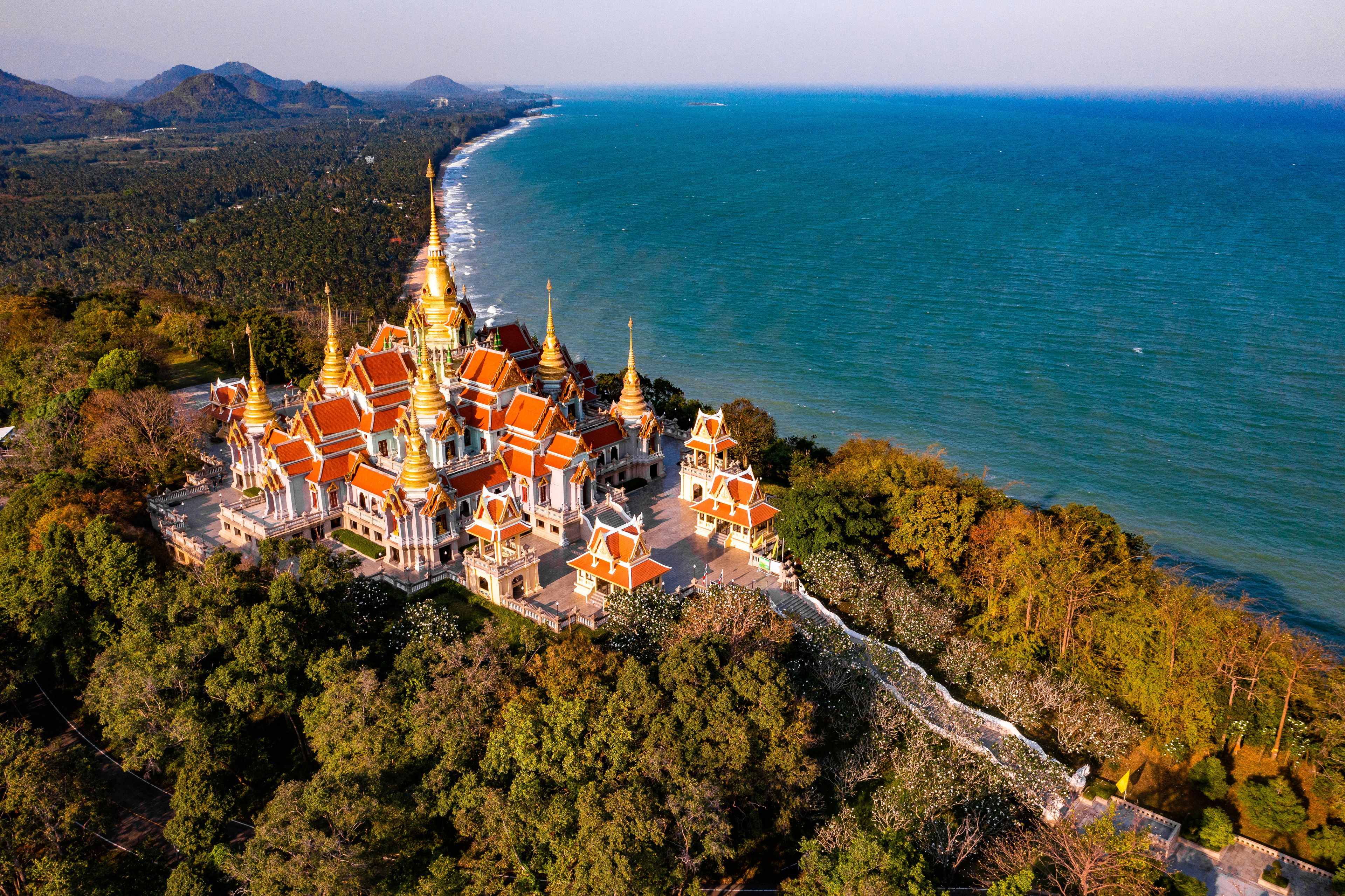 An aerial view of a temple complex with golden spires, surrounded by lush greenery and the coastline.