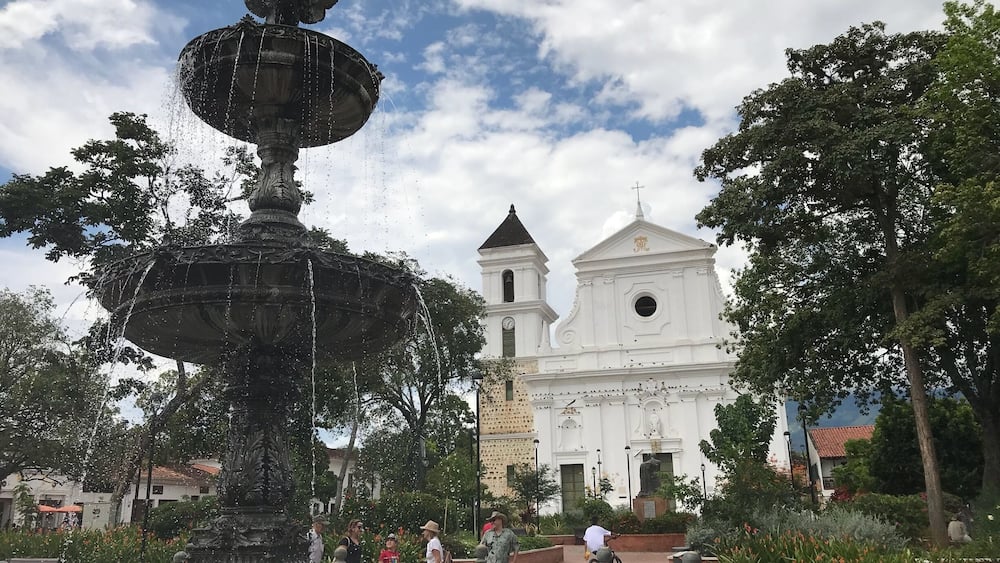 Une fontaine historique, agrémentée de jets d’eau, entourée d’une végétation luxuriante et, en arrière-plan, une église blanche dotée d’un clocher.
