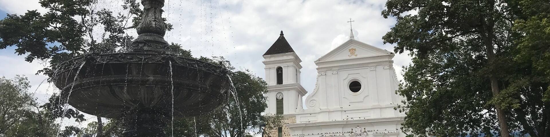 A historic fountain with water jets, surrounded by lush greenery and a white church with a bell tower in the background.