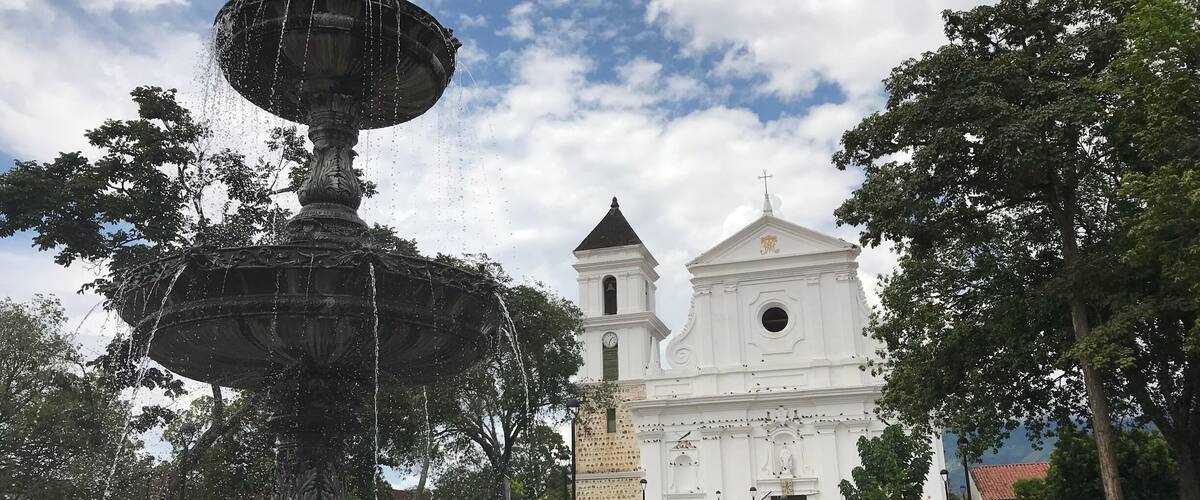 A historic fountain with water jets, surrounded by lush greenery and a white church with a bell tower in the background.
