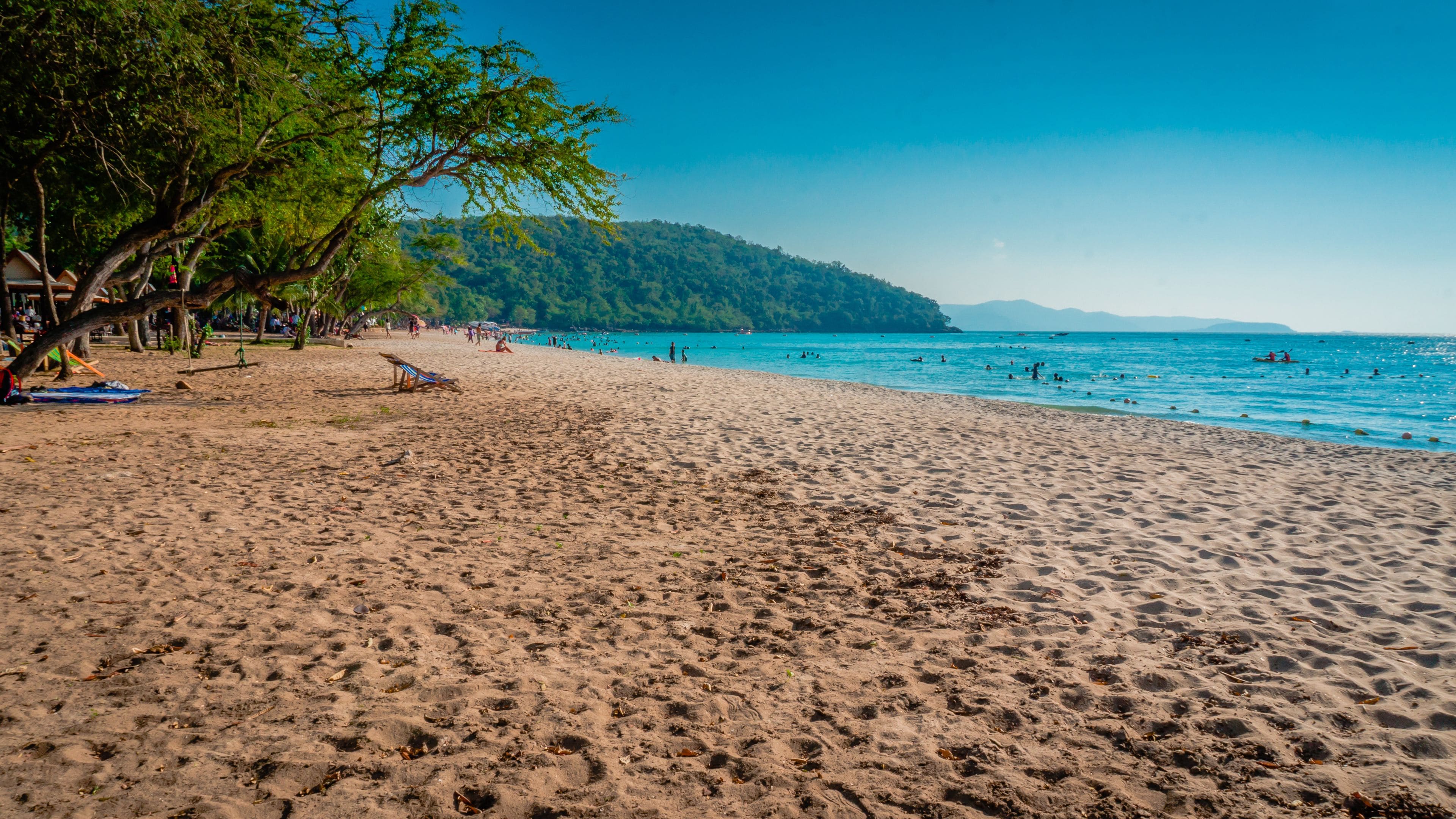 Uma praia arenosa com pessoas nadando e tomando sol, céu azul límpido e uma colina arborizada ao fundo.