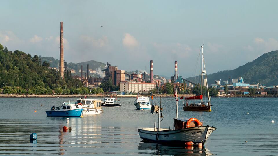 A harbor with multiple boats, industrial buildings, and smokestacks in the background.