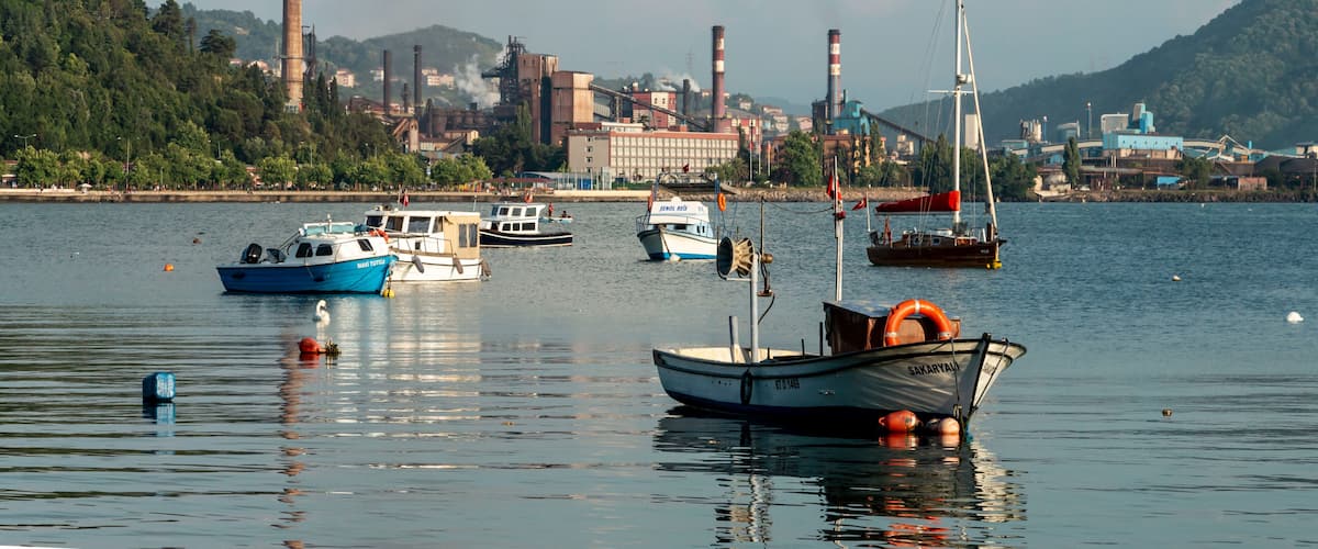A harbor with multiple boats, industrial buildings, and smokestacks in the background.