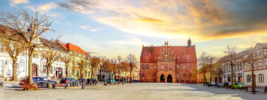 A cobblestone square with historic buildings, a prominent red brick structure, and leafless trees.