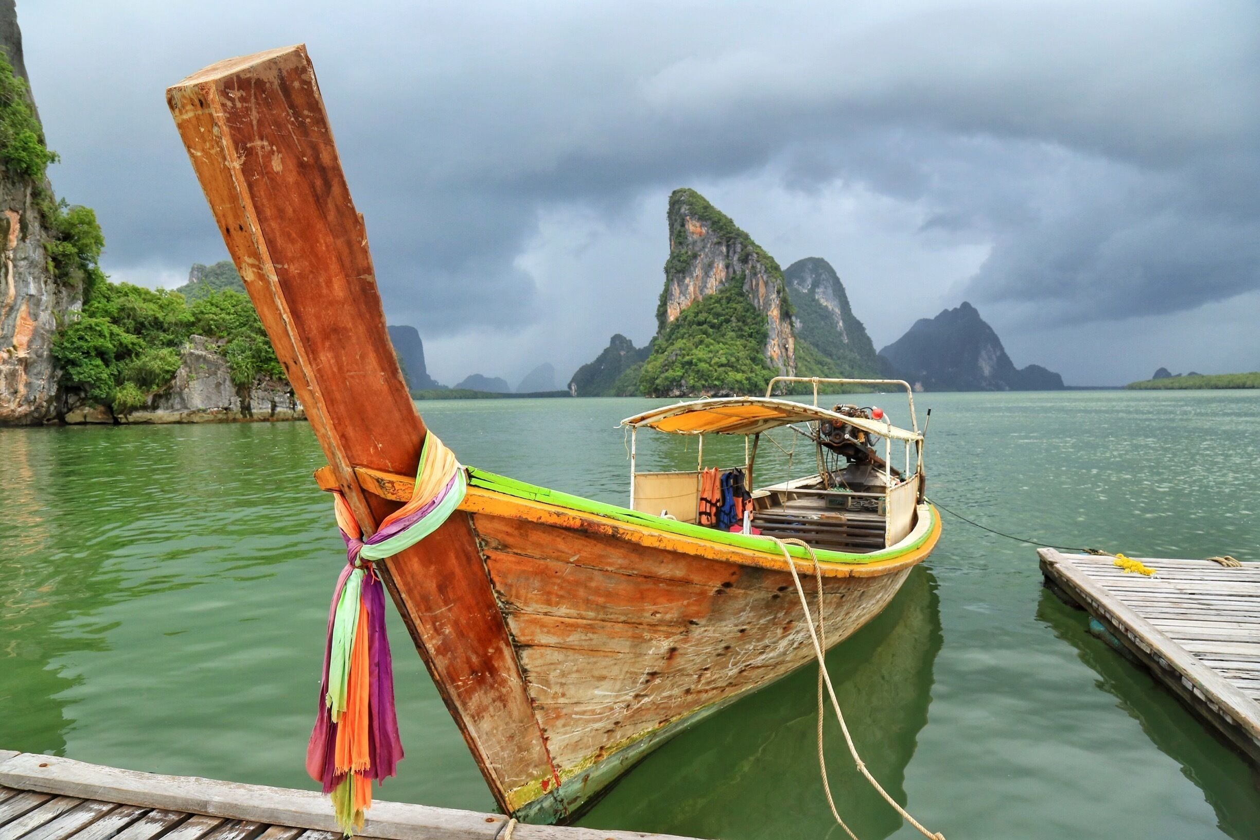 A traditional wooden boat tied to a dock with a backdrop of green cliffs and overcast sky.