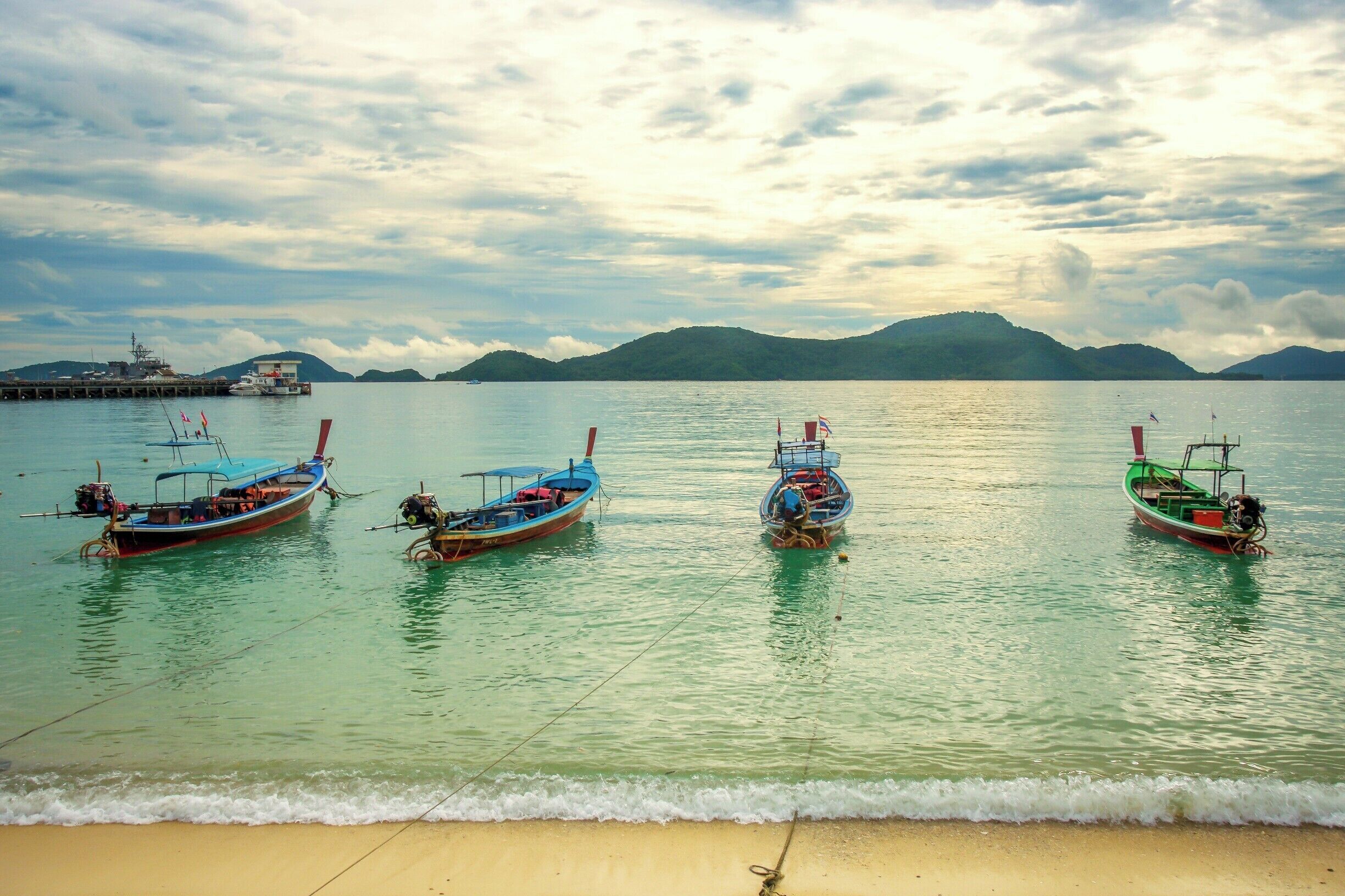 A calm sea with several boats anchored near a sandy beach and a pier in the background.