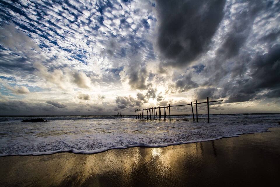 A beach with a wooden pier extending into the ocean, waves gently lapping at the shore, and a dramatic sky with clouds.