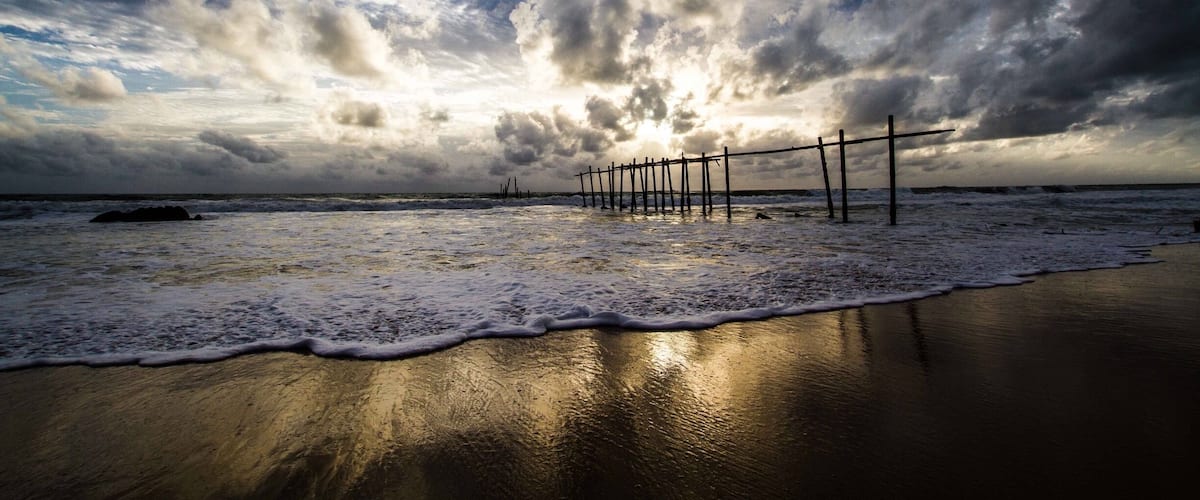 A beach with a wooden pier extending into the ocean, waves gently lapping at the shore, and a dramatic sky with clouds.