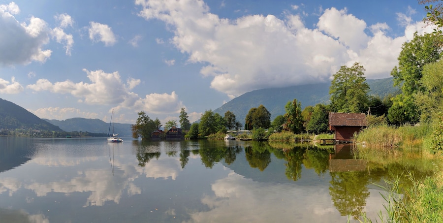 A calm lake with reflections of trees and a sailboat, surrounded by mountains and houses.