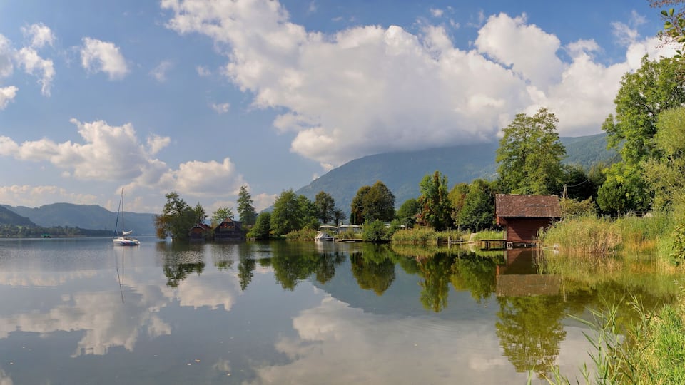 A calm lake with reflections of trees and a sailboat, surrounded by mountains and houses.