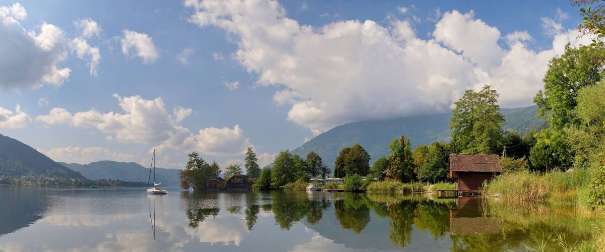 A calm lake with reflections of trees and a sailboat, surrounded by mountains and houses.