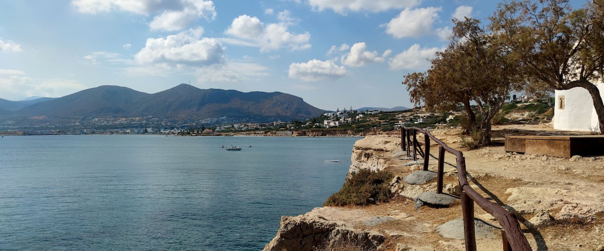 A coastal landscape with a rocky shoreline, a wooden fence, a white building, and mountains in the background.